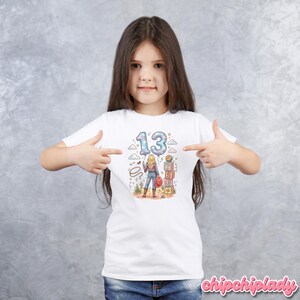 Pu&ograve; includere: Una t-shirt bianca a maniche corte con una stampa grafica di una persona in piedi in un paesaggio desertico. Il design include il numero 13 a forma di palloncino, un lazo, un cappello rosso e blocchi impilati con le lettere T, S ed E stampate.