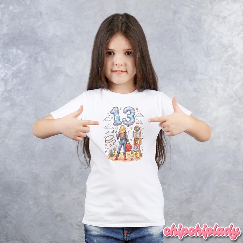 Pu&ograve; includere: Una t-shirt bianca a maniche corte con una stampa grafica di una persona in piedi in un paesaggio desertico. Il design include il numero 13 a forma di palloncino, un lazo, un cappello rosso e blocchi impilati con le lettere T, S ed E stampate.