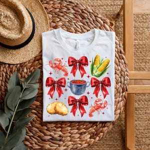 May include: White t-shirt with a red, white, and yellow design featuring a pot of gumbo, crawfish, corn on the cob, and red bows.