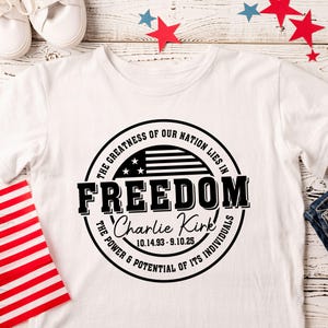 Peut inclure: T-shirt blanc avec un motif circulaire noir portant l'inscription "FREEDOM" et le texte "La grandeur de notre nation r&eacute;side dans..." et "Le pouvoir et le potentiel de ses individus". Comprend un drapeau am&eacute;ricain et le nom "Charlie Kirk".