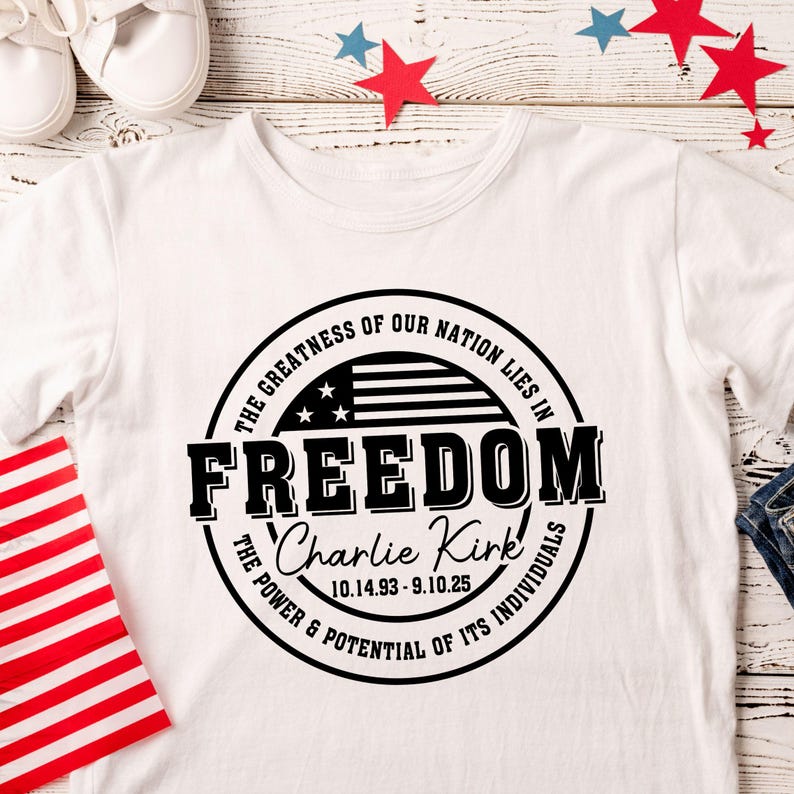 Peut inclure: T-shirt blanc avec un motif circulaire noir portant l'inscription "FREEDOM" et le texte "La grandeur de notre nation r&eacute;side dans..." et "Le pouvoir et le potentiel de ses individus". Comprend un drapeau am&eacute;ricain et le nom "Charlie Kirk".