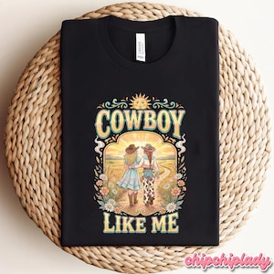 May include: A black t-shirt featuring a graphic print of two figures in cowboy hats walking down a dirt path toward a sun. The text COWBOY LIKE ME is printed in yellow above and below the illustration, surrounded by colorful flowers and foliage.
