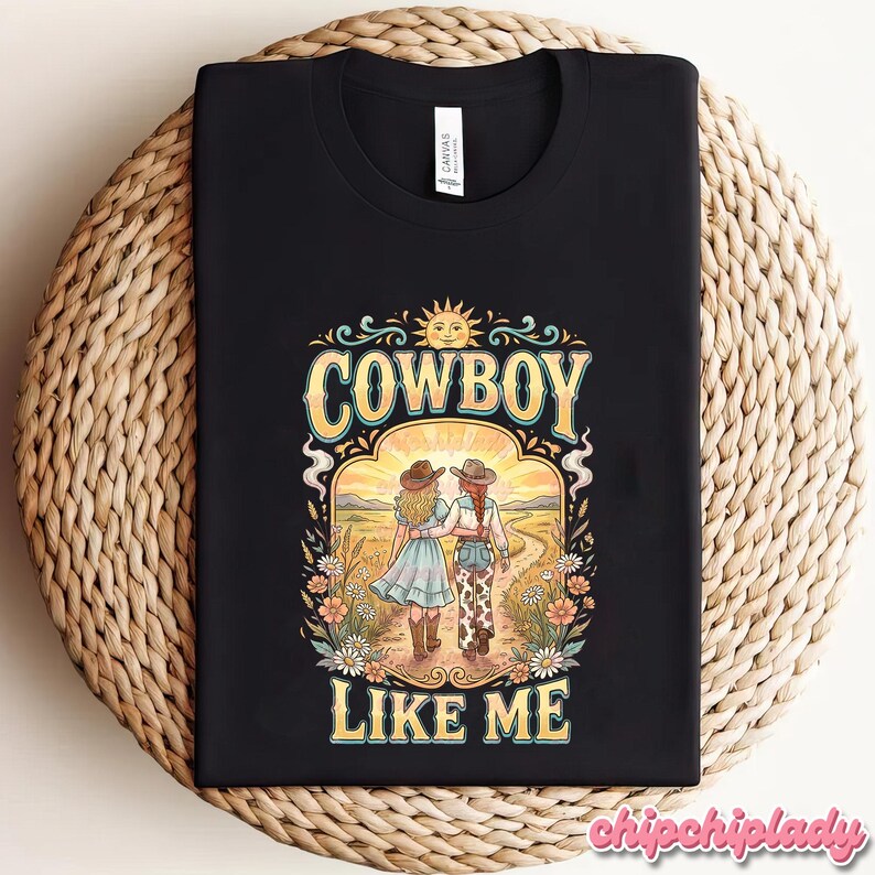 May include: A black t-shirt featuring a graphic print of two figures in cowboy hats walking down a dirt path toward a sun. The text COWBOY LIKE ME is printed in yellow above and below the illustration, surrounded by colorful flowers and foliage.
