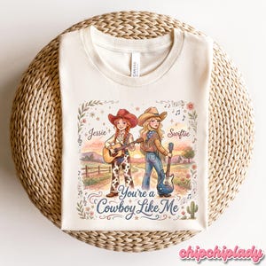 Pu&ograve; includere: Una t-shirt color crema con una grafica di due cowgirl in un paesaggio con il testo Jessie, Swiftie e You're a Cowboy Like Me. L'illustrazione include note musicali, fiori, un cactus e chitarre in una delicata tavolozza di colori.