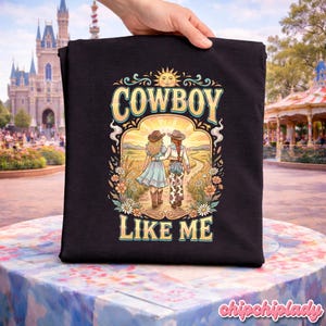 May include: A black t-shirt featuring a graphic print of two individuals in cowboy hats walking away on a dirt path. The text COWBOY LIKE ME is printed in yellow. The illustration includes a sun, flowers, and a landscape background.