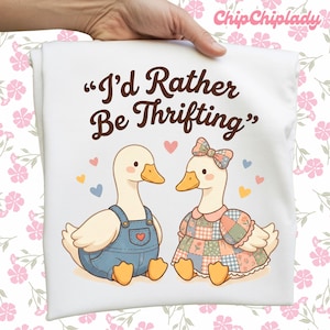 May include: White t-shirt featuring a cartoon illustration of two ducks. One duck wears blue overalls, the other a patchwork dress. The text "I'd Rather Be Thrifting" is printed above the ducks, with hearts scattered around.