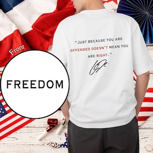 Peut inclure: T-shirt blanc avec le texte "JUST BECAUSE YOU ARE OFFENDED DOESN'T MEAN YOU ARE RIGHT." en noir et rouge. Le mot "FREEDOM" est imprim&eacute; sur un cercle blanc. Le t-shirt est entour&eacute; de drapeaux am&eacute;ricains et fran&ccedil;ais.