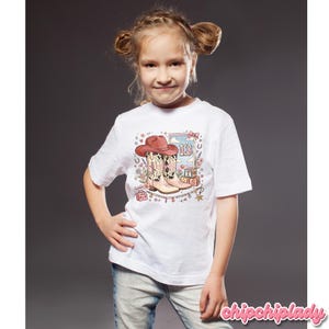 Pode incluir: Uma t-shirt branca de manga curta com uma estampa gr&aacute;fica de um chap&eacute;u de cowboy rosa, um par de botas de cowboy com estampa de vaca, o n&uacute;mero 13 e blocos alfab&eacute;ticos que formam T-S. O design inclui detalhes florais, estrelas e cora&ccedil;&otilde;es.