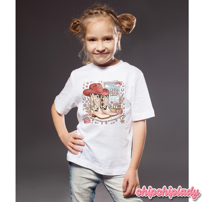 Pode incluir: Uma t-shirt branca de manga curta com uma estampa gr&aacute;fica de um chap&eacute;u de cowboy rosa, um par de botas de cowboy com estampa de vaca, o n&uacute;mero 13 e blocos alfab&eacute;ticos que formam T-S. O design inclui detalhes florais, estrelas e cora&ccedil;&otilde;es.
