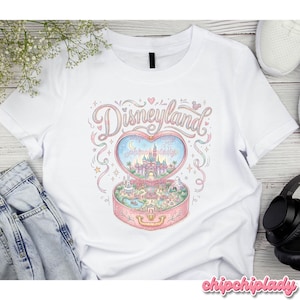 Może przedstawiać: Biała koszulka z pastelową ilustracją Disneylandu wewnątrz kompaktowego pudełka w kształcie serca. Projekt zawiera napis "Disneyland" w kursywie, gwiazdy, fajerwerki i zamek. Koszulka wykonana jest z miękkiego materiału.