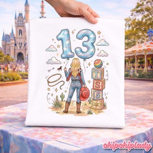 Pu&ograve; includere: Una t-shirt bianca con un'illustrazione grafica di una persona bionda in tenuta da cowgirl, che tiene due grandi palloncini a forma di numero 13. Il disegno include un lazo, cactus e una pila di blocchi con le lettere T, S e il numero 5.