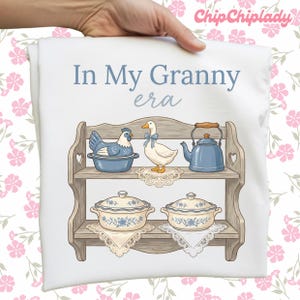 May include: White pillowcase with the text "In My Granny era" and a graphic of a wooden shelf with a blue chicken, a white duck with a blue bow, a blue teapot, and two blue and white casserole dishes.