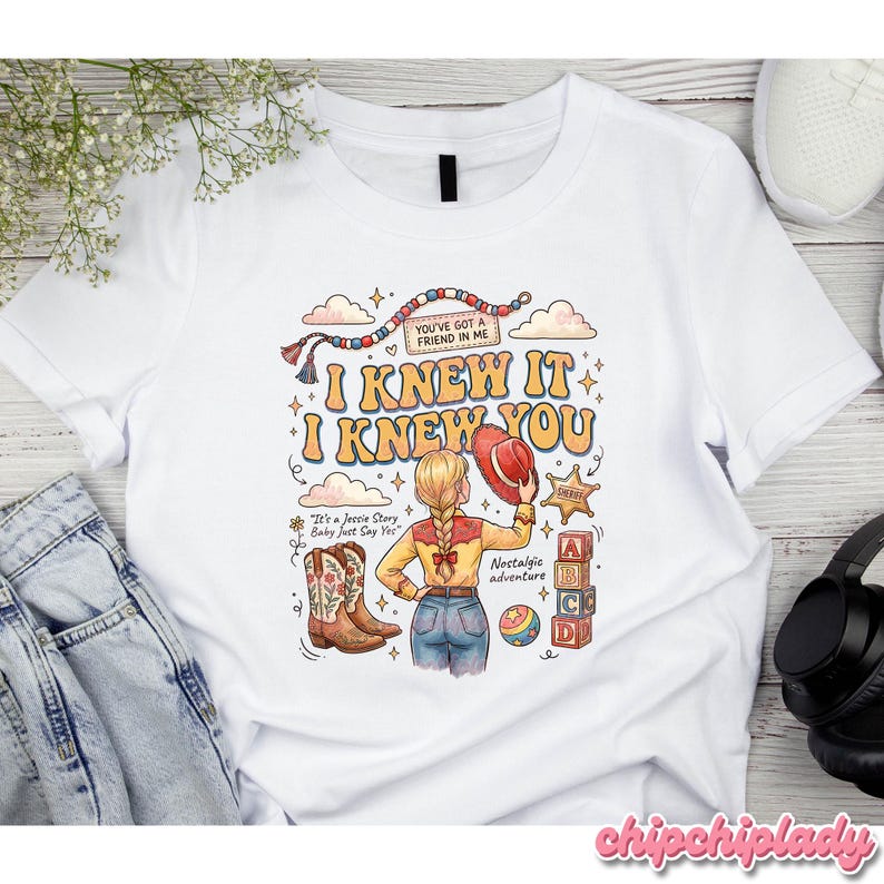 May include: A white t-shirt featuring a graphic illustration of a blonde character in a western outfit, cowboy boots, and a hat. The design includes text that reads "I KNEW IT I KNEW YOU", "YOU'VE GOT A FRIEND IN ME", "It's a Jessie Story Baby Just Say Yes", and "Nostalgic adventure" with alphabet blocks.