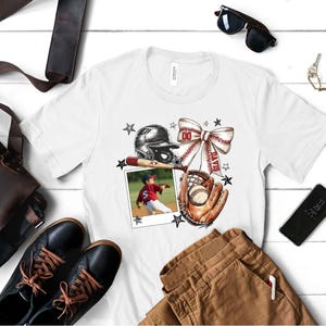 May include: White t-shirt with a baseball-themed design. The design includes a baseball glove, bat, ball, helmet, and a bow with the word "DAVE's" on it. A photo of a young baseball player is in the center of the design.