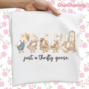 May include: White t-shirt featuring a whimsical illustration of six geese dressed in various outfits, including overalls, dresses, and accessories. The text "just a thrifty goose" is printed below the geese.
