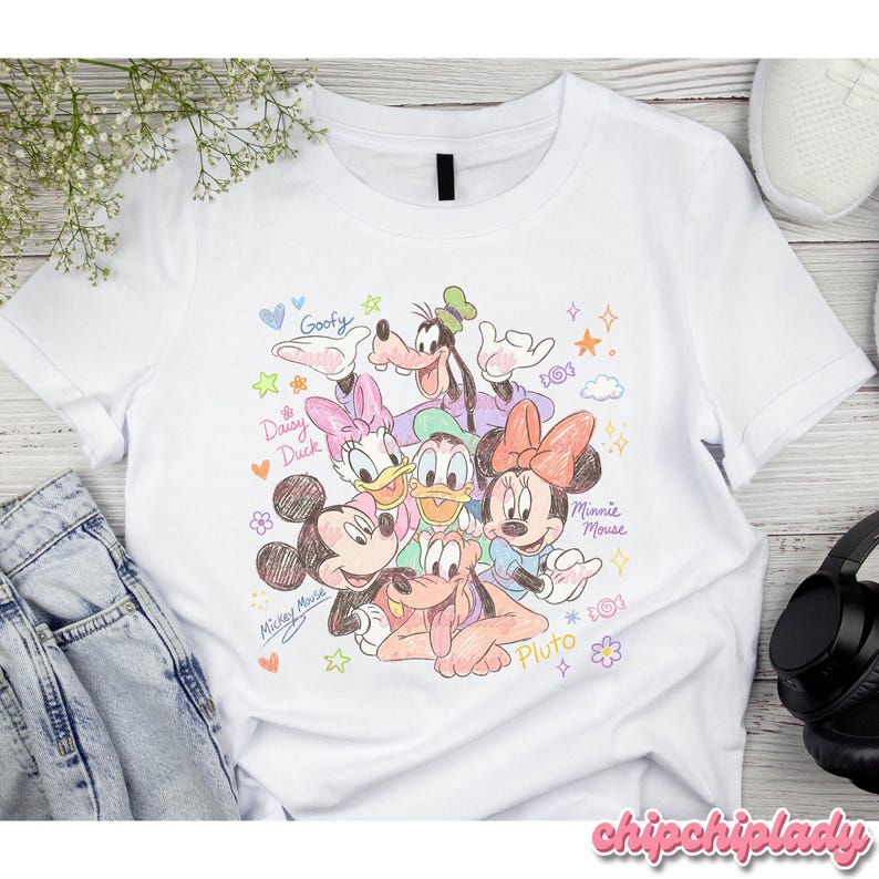 May include: White t-shirt featuring a colorful illustration of Disney characters Mickey Mouse, Minnie Mouse, Donald Duck, Daisy Duck, Goofy, and Pluto. The design includes the characters' names and decorative elements like hearts and stars.