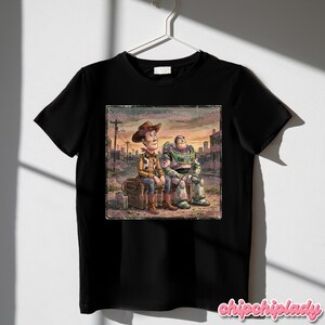 May include: Black t-shirt featuring a distressed graphic of Woody and Buzz Lightyear from Toy Story, sitting together in a post-apocalyptic setting. The image has a vintage, watercolor style. The shirt is hanging on a white hanger.