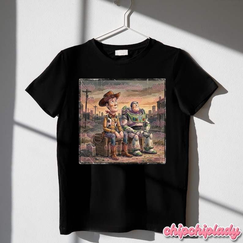 May include: Black t-shirt featuring a distressed graphic of Woody and Buzz Lightyear from Toy Story, sitting together in a post-apocalyptic setting. The image has a vintage, watercolor style. The shirt is hanging on a white hanger.