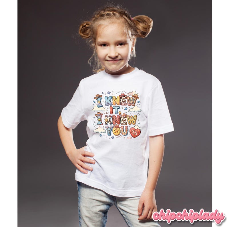 K&ouml;nnte beinhalten: Ein wei&szlig;es T-Shirt mit kurzen &Auml;rmeln und einem bunten Grafikdruck mit dem Text I KNEW IT, I KNEW YOU. Das Design enth&auml;lt Cowboyh&uuml;te, Sterne, Wolken und eine Herzform mit den Buchstaben TS, alles in verschiedenen Mustern und Farben dargestellt.