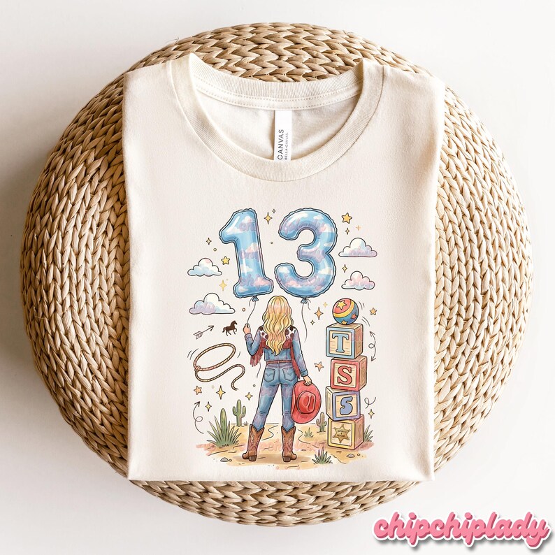Pu&ograve; includere: Una t-shirt color crema con un'illustrazione grafica di una persona vista di spalle, che tiene due palloncini blu con motivo a nuvole a forma di numero 13. Il disegno include un lazo, una pila di blocchi con le lettere T, S, S e una stella.