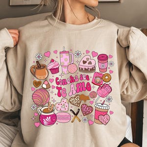 Op de afbeelding: Beige sweatshirt met een kleurrijke afbeelding van diverse zoetigheden en drankjes, waaronder cupcakes, koffiebekers en koekjes. De tekst "Concha see Te Amo" staat in het midden, omringd door hartjes en bloemen. Een vrolijk en feestelijk ontwerp.