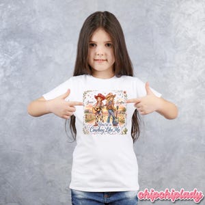 Pu&ograve; includere: Una t-shirt bianca con un'illustrazione grafica di due cowgirl con chitarre in un paesaggio rurale. Il testo recita Jessie, Swiftie e You're a Cowboy Like Me, circondato da accenti floreali e un cactus. Il disegno &egrave; centrato sul petto.