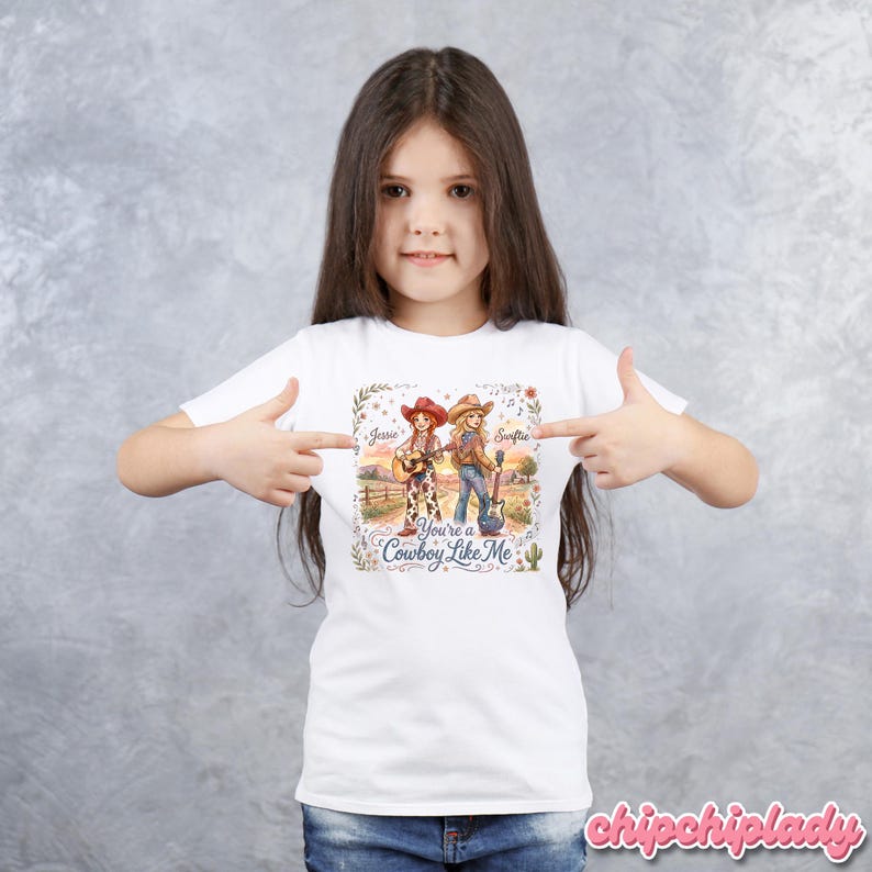 Pu&ograve; includere: Una t-shirt bianca con un'illustrazione grafica di due cowgirl con chitarre in un paesaggio rurale. Il testo recita Jessie, Swiftie e You're a Cowboy Like Me, circondato da accenti floreali e un cactus. Il disegno &egrave; centrato sul petto.