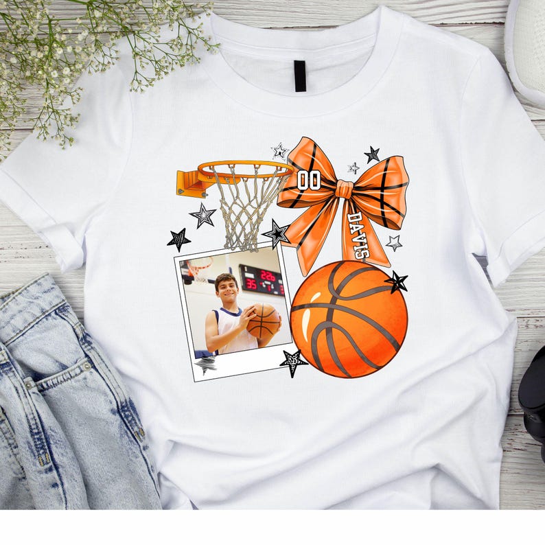 May include: White t-shirt with a basketball design. The design includes a basketball hoop, a basketball, a bow with the name "Davis" and the number "00" on it, and a photo of a young person holding a basketball. The design is surrounded by stars.