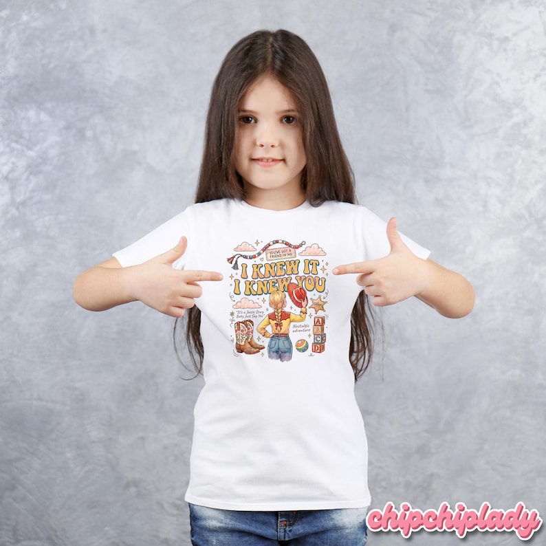 May include: A white short-sleeved t-shirt featuring a colourful graphic print. The design includes the text I KNEW IT I KNEW YOU, a blonde character wearing a hat, cowboy boots, alphabet blocks, and a ball, all surrounded by decorative stars and clouds.