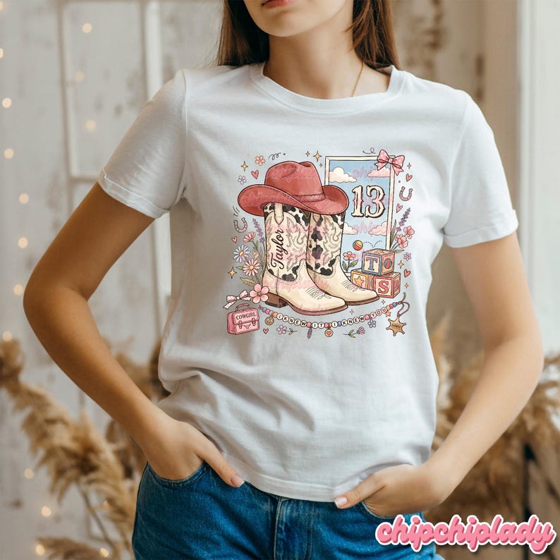 Pode incluir: Uma t-shirt branca de manga curta com uma impress&atilde;o gr&aacute;fica de um chap&eacute;u de cowboy rosa sobre um par de botas western. O design inclui o nome Taylor, o n&uacute;mero 13, blocos de letras, flores, estrelas e uma pequena mala cor-de-rosa.