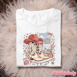 Pode incluir: Uma t-shirt branca com uma ilustra&ccedil;&atilde;o gr&aacute;fica de um chap&eacute;u de cowboy rosa sobre um par de botas western com padr&atilde;o de vaca. O design inclui o nome Taylor, o n&uacute;mero 13, blocos de alfabeto, flores e o texto I KNEW IT I KNEW YOU.
