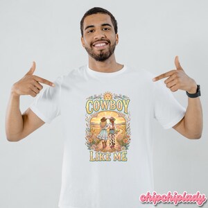 May include: A white short-sleeved t-shirt featuring a colorful graphic print of two figures in cowboy hats walking down a path. The design includes floral elements, a sun, and the text "COWBOY LIKE ME" in a stylized, retro-inspired font.