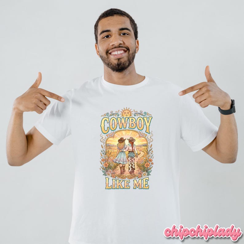 May include: A white short-sleeved t-shirt featuring a colorful graphic print of two figures in cowboy hats walking down a path. The design includes floral elements, a sun, and the text "COWBOY LIKE ME" in a stylized, retro-inspired font.