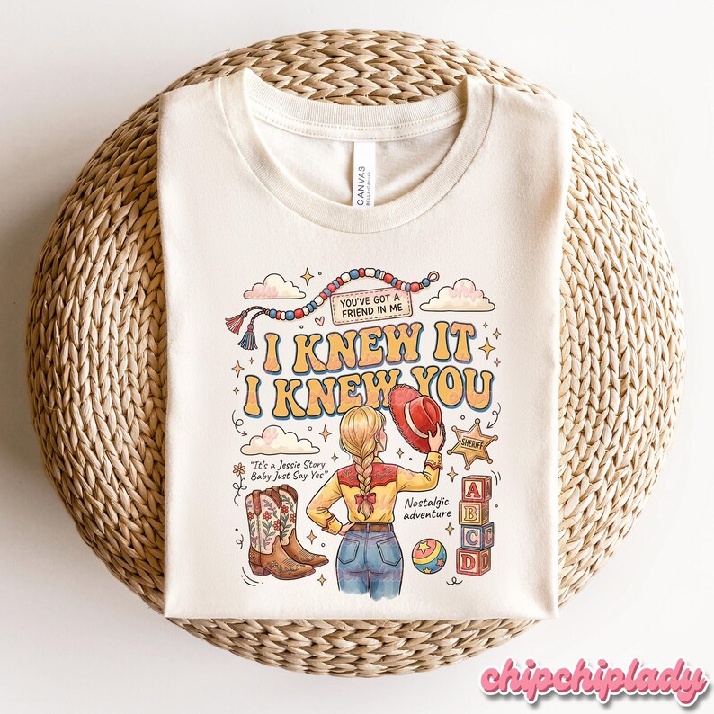 May include: A cream-coloured t-shirt featuring a graphic illustration of a person with blonde braided hair wearing a yellow shirt and blue jeans, holding a red hat. The design includes cowboy boots, a sheriff badge, alphabet blocks, and text.
