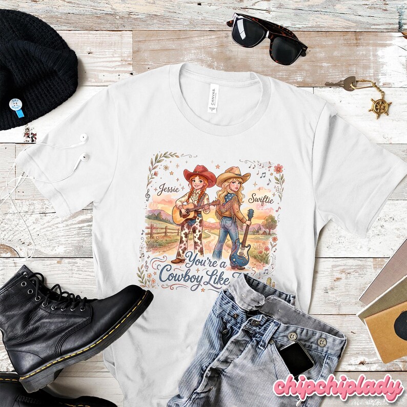 Pu&ograve; includere: Una t-shirt bianca a maniche corte con un'illustrazione grafica di due cowgirl con chitarre in un paesaggio rurale. Il testo Jessie e Swiftie appare sopra le figure, con la frase You're a Cowboy Like Me stampata in basso.