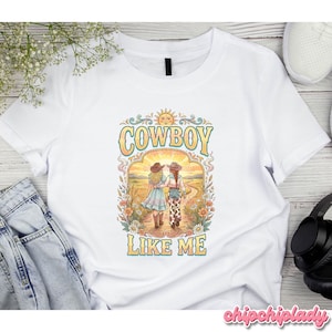 May include: A white t-shirt featuring a graphic print of two people walking away on a dirt path through a field. The design includes floral borders, a sun, and the text "COWBOY LIKE ME" in a stylized yellow font with a blue outline.