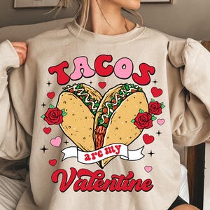 May include: A beige sweatshirt with a graphic design of two tacos in the shape of a heart. The text "Tacos are my Valentine" is printed in red and pink letters.