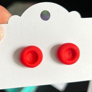 May include: A pair of red stud earrings with a slightly raised center. The earrings are displayed on a white card with a scalloped edge.