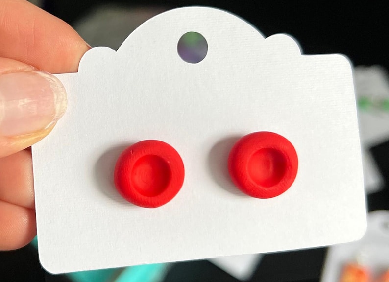 May include: A pair of red stud earrings with a slightly raised center. The earrings are displayed on a white card with a scalloped edge.