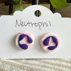 May include: A pair of purple and pink swirl polymer clay earrings with the word "Neutrophil" written on a white card.