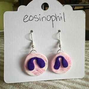 May include: A pair of earrings with a pink and white marbled design and a purple shape on each earring. The earrings are on a white card with the word "eosinophil" written on it.