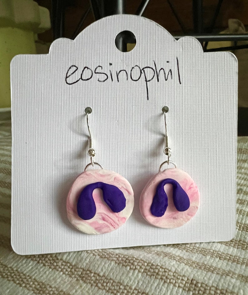 May include: A pair of earrings with a pink and white marbled design and a purple shape on each earring. The earrings are on a white card with the word "eosinophil" written on it.