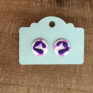 May include: A pair of white and purple swirl stud earrings. The earrings are round and have a white background with purple swirls.