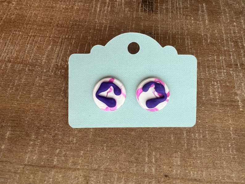 May include: A pair of white and purple swirl stud earrings. The earrings are round and have a white background with purple swirls.