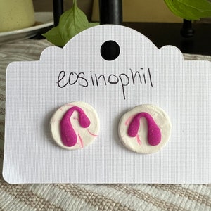 May include: A pair of pink and white clay earrings with a unique shape. The earrings are on a white card with the text "eosinophil" written on it.