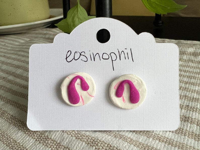 May include: A pair of pink and white clay earrings with a unique shape. The earrings are on a white card with the text "eosinophil" written on it.