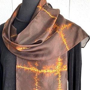 May include: A brown silk scarf with a tie-dye pattern in shades of orange and yellow. The scarf is folded and draped around a mannequin's neck.