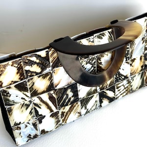Può includere: Una pochette rettangolare con un motivo a mosaico di quadrati bianco sporco, neri e marroni. La borsa è dotata di un manico semicircolare marrone scuro. I bordi della borsa sono neri.