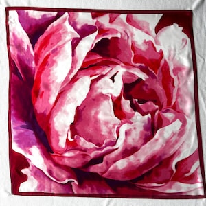 May include: Square silk scarf featuring a close-up of a pink and red peony flower. The floral design has white highlights and a dark red border. The scarf is likely made of a soft, flowing material.