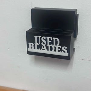 May include: A black wall-mounted disposal container for razor blades featuring a rectangular shape and a top slot. The front of the container displays the white embossed text "USED BLADES" in a bold, sans-serif font, clearly indicating its function.